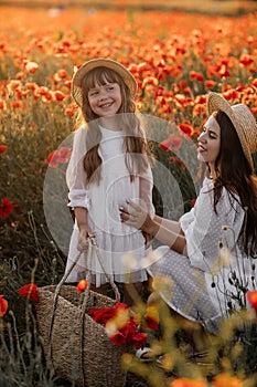 Beautiful young woman with girl in field with poppies, mother and daughter in white dresses and straw hats in evening at sunset,