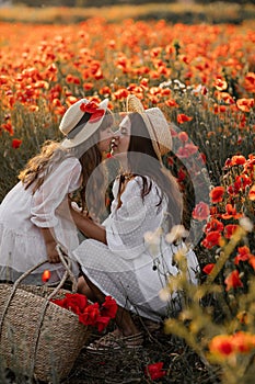 Beautiful young woman with girl in field with poppies, mother and daughter in white dresses and straw hats in evening at sunset,