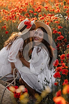 Beautiful young woman with girl in field with poppies, mother and daughter in white dresses and straw hats in evening at sunset,