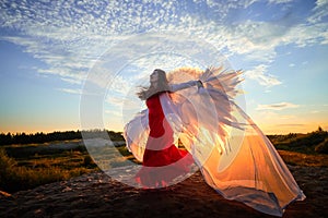 Beautiful young woman or girl in red dress and white wings on the sand on sunny day with blue sky. Angel model or dancer