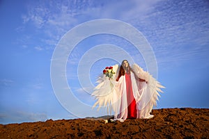 Beautiful young woman or girl in red dress and white wings on the sand on sunny day with blue sky. Angel model or dancer