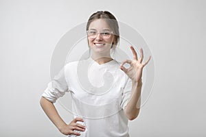 Beautiful young woman in glasses is looking at camera, showing Ok sign and smiling.