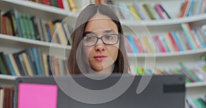 Beautiful young woman in glasses reading book and typing on computer. College student works on essay at the library.