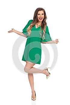 Beautiful Young Woman In Green Dress And High Heels Is Standing On One Leg And Laughing With Arms Outstretched