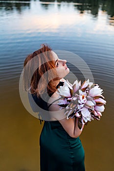 Beautiful young woman in green silk evening dress holding water lily flower outdoors.