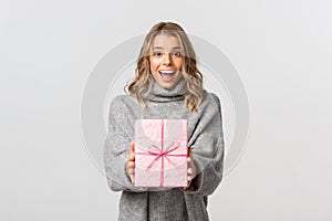 Beautiful young woman in grey sweater, giving gift, congratulating someone with birthday, standing over white background