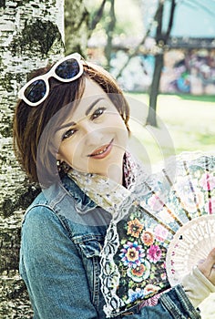 Beautiful young woman with hand fan is leaning on the birch tree