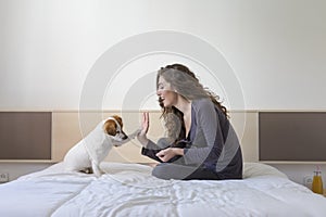 beautiful young woman hand high five with cute small dog over white background. Dog is sitting on bed. Daytime, pets indoors,