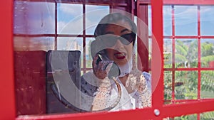 Beautiful young woman happily speaks on the phone in an English style red telephone booth. Girl dressed in a white dress