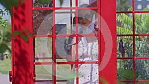 Beautiful young woman happily speaks on the phone in an English style red telephone booth. Girl dressed in a white dress