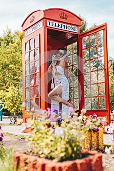 beautiful young woman happily speaks on the phone in an english style red telephone booth. girl dressed in a white dress