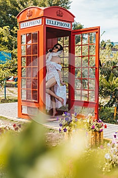 beautiful young woman happily speaks on the phone in an english style red telephone booth. girl dressed in a white dress