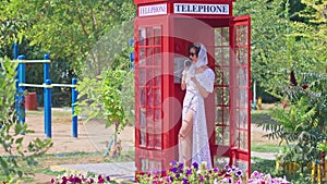 Beautiful young woman happily speaks on the phone in an English style red telephone booth. Girl dressed in a white dress
