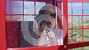 beautiful young woman happily speaks on the phone in an english style red telephone booth. girl dressed in a white dress