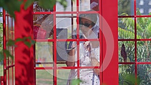 beautiful young woman happily speaks on the phone in an english style red telephone booth. girl dressed in a white dress