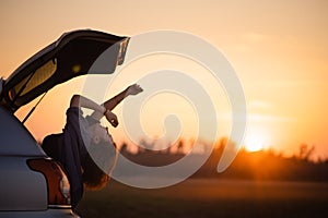 Beautiful young woman happy and dancing in a car`s trunk during a road trip in Europe in the last minutes of Golden Hour