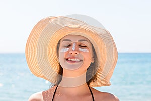 Beautiful young woman in hat is applying sunblock under her eyes and on her nose like Indian. Sun protection concept