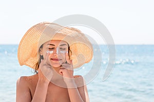 Beautiful young woman in hat is applying sunblock under her eyes and on her nose like Indian. Sun protection concept