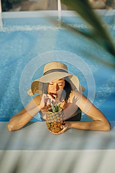Beautiful young woman in hat drinking delicious cocktail from pineapple and relaxing in pool, summer vacation. Girl enjoying warm