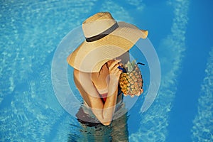 Beautiful young woman in hat drinking delicious cocktail from pineapple and relaxing in pool, summer vacation. Girl enjoying warm