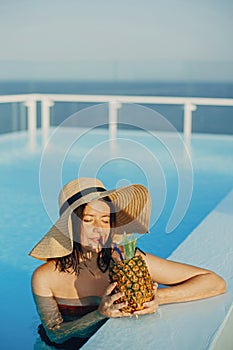 Beautiful young woman in hat drinking delicious cocktail from pineapple and relaxing in pool, summer vacation. Girl enjoying warm