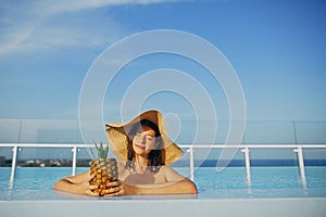 Beautiful young woman in hat holding delicious cocktail in pineapple and relaxing in pool. Summer vacation. Girl enjoying warm