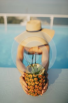 Beautiful young woman in hat holding delicious cocktail in pineapple and relaxing in pool, summer vacation. Girl enjoying warm