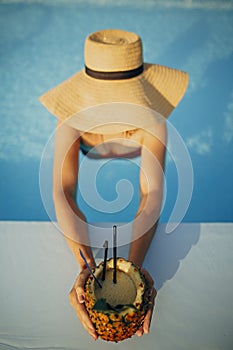 Beautiful young woman in hat holding delicious cocktail in pineapple and relaxing in pool, summer vacation. Girl enjoying warm