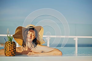 Beautiful young woman in hat relaxing in pool with delicious cocktail in pineapple. Summer vacation. Girl enjoying warm sunshine