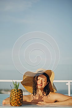 Beautiful young woman in hat relaxing in pool with delicious cocktail in pineapple. Summer vacation. Girl enjoying warm sunshine