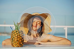 Beautiful young woman in hat relaxing in pool with delicious cocktail in pineapple. Summer vacation. Girl enjoying warm sunshine