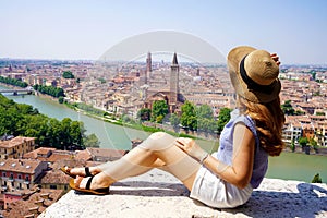 Beautiful young woman with hat sitting on wall looking at stunning panoramic view of Verona City with Adige River, Italy
