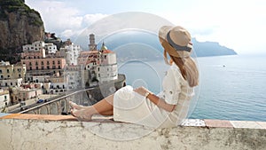 Beautiful young woman with hat sitting on wall looking at stunning panoramic village of Atrani on Amalfi Coast, Italy