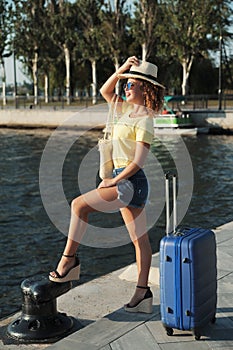 Beautiful young woman in hat with suitcase and wine glass waiting a ship for summer trip.