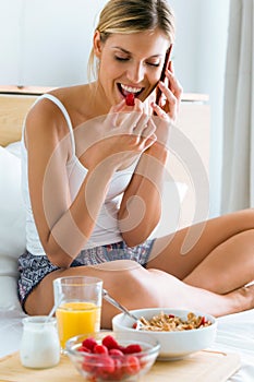 Beautiful young woman having breakfast and talking with her smartphone in the bedroom.