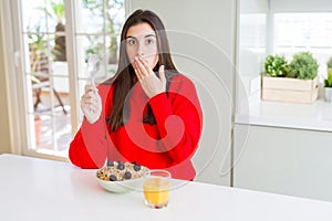 Beautiful young woman having healthy cereals and berries for breakfast cover mouth with hand shocked with shame for mistake,