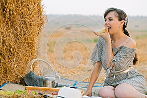 Beautiful young woman having a picnic and eats some bread near a hay bale in the middle of a wheat field in summer