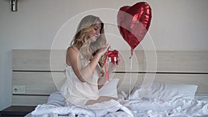 A beautiful young woman with a heart-shaped balloon sits on the bed at home holds a red box with a bow in her hands and