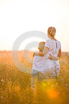 Beautiful young woman with her son on the daisy meadow on a sunny day. Happy family on summer sunset. Kid boy hugging his mother.