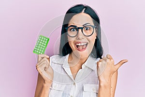 Beautiful young woman holding birth control pills pointing thumb up to the side smiling happy with open mouth