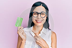 Beautiful young woman holding birth control pills smiling happy pointing with hand and finger