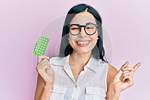 Beautiful young woman holding birth control pills smiling happy pointing with hand and finger to the side