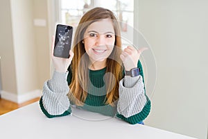 Beautiful young woman holding broken smartphone pointing and showing with thumb up to the side with happy face smiling