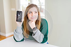 Beautiful young woman holding broken smartphone serious face thinking about question, very confused idea