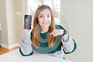 Beautiful young woman holding broken smartphone with surprise face pointing finger to himself