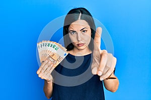 Beautiful young woman holding bunch of 50 euro banknotes pointing with finger up and angry expression, showing no gesture