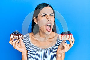 Beautiful young woman holding cake slices angry and mad screaming frustrated and furious, shouting with anger