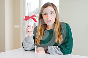 Beautiful young woman holding degree with red bow scared in shock with a surprise face, afraid and excited with fear expression