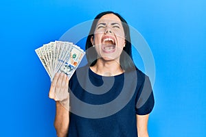 Beautiful young woman holding dollars angry and mad screaming frustrated and furious, shouting with anger