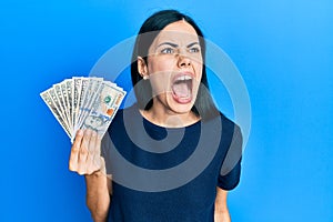 Beautiful young woman holding dollars angry and mad screaming frustrated and furious, shouting with anger